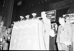Eddie Cantor standing at a podium speaking with people in background