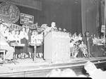 Eddie Cantor standing at a podium speaking, people in background
